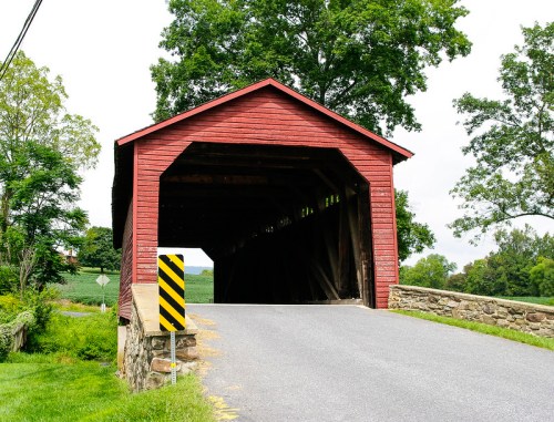 A red covered bridge with a stone foundation, leading to a rural road surrounded by greenery.