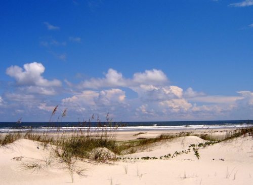 A serene beach scene with soft white sand, gentle waves, and a blue sky dotted with fluffy clouds.