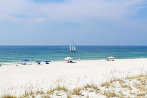 A serene beach scene with white sand, blue umbrellas, and a sailboat on calm waters under a clear sky.