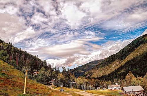A scenic view of mountains with lush greenery, a winding road, and a ski lift under a cloudy blue sky.
