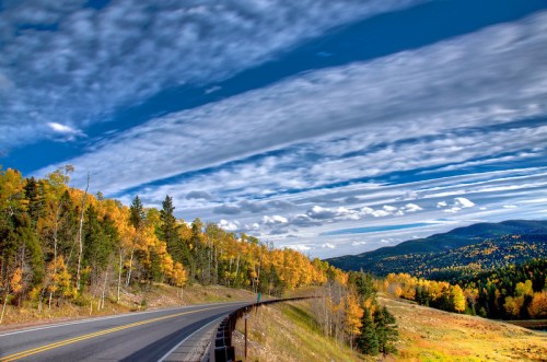 A winding road through vibrant autumn trees under a blue sky with wispy clouds.