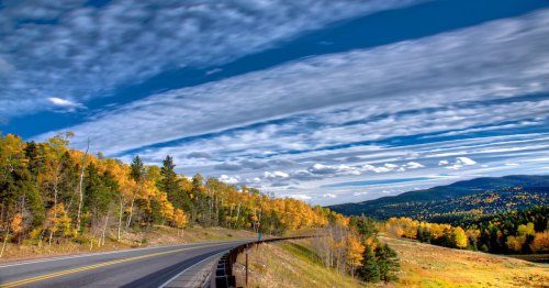 A winding road through vibrant autumn trees under a blue sky with wispy clouds and distant mountains.