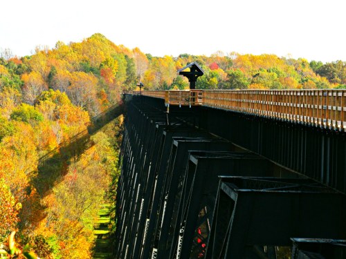 A tall bridge spans a colorful autumn landscape, with vibrant trees and long shadows cast on the ground below.