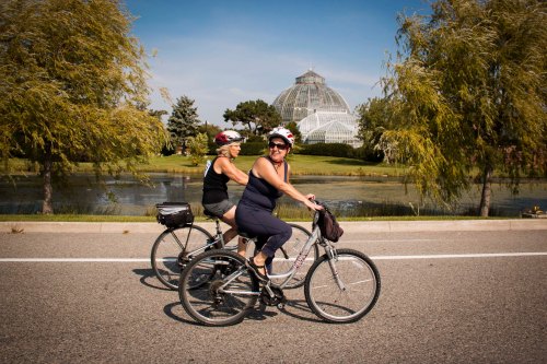 Two women ride bicycles along a road, with a glass conservatory and trees in the background under a clear blue sky.