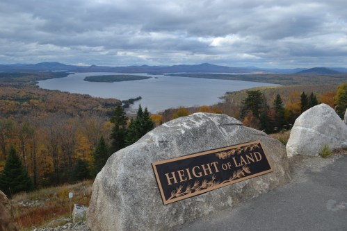 View from Height of Land, showcasing a lake surrounded by autumn foliage and distant mountains under a cloudy sky.