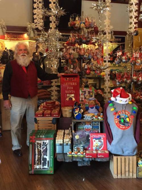 A man in a red vest stands beside a festive display of holiday decorations and gifts in a cozy shop.