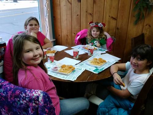 Four children sit at a wooden table, enjoying snacks and drinks in a cozy setting.