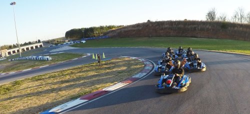 A group of go-karts racing on a track, with a clear sky and green grass in the background.
