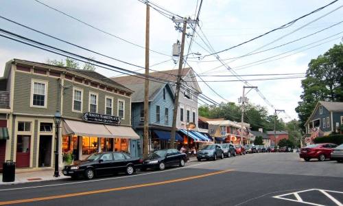 A quaint street lined with shops and parked cars, featuring power lines and a cloudy sky overhead.
