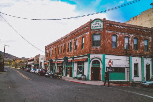 Historic brick buildings line a street, featuring Hotel Connor and various shops, with mountains in the background.