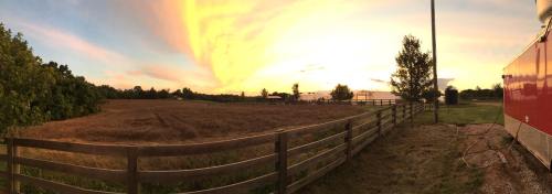 A panoramic view of a sunset over a field, with trees and a barn in the distance, framed by a wooden fence.