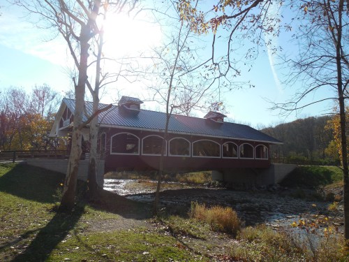 A red covered bridge spans a stream, surrounded by autumn foliage and sunlight filtering through trees.