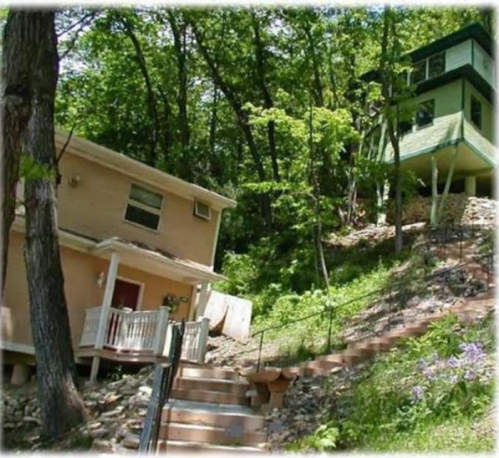 A hillside scene featuring two houses: a beige house on the left and a green multi-level house on the right, surrounded by trees.