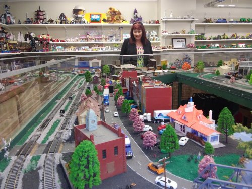 A woman stands behind a display of a miniature town with buildings, trees, and train tracks in a model exhibit.