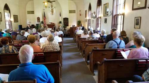 A congregation seated in a church, facing the front where a service is taking place, with flags and plants in the background.