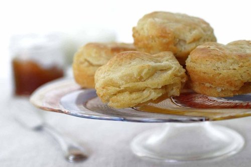 A plate of golden, flaky biscuits with a jar of jam in the background.