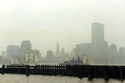 Foggy skyline with tall buildings and ships in the harbor, viewed from a wooden pier.