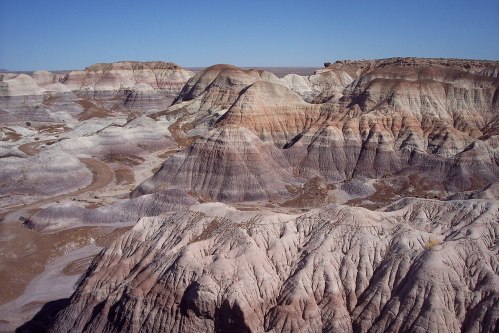 A vast landscape of colorful, layered hills and valleys under a clear blue sky, showcasing unique geological formations.