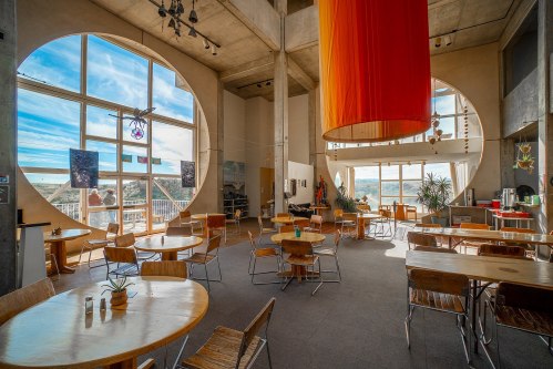 Bright, modern café with large windows, circular design, wooden tables, and colorful hanging fabric.