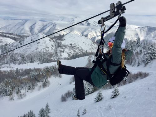 A person zip-lining over a snowy landscape with mountains and trees in the background.