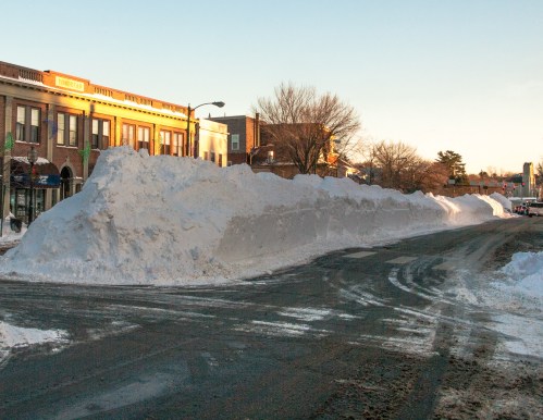 A large snowbank lines the side of a street, with buildings visible in the background under a clear sky.