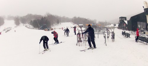 Skiers preparing on a snowy slope with a ski lift and lodge in the background, surrounded by winter scenery.
