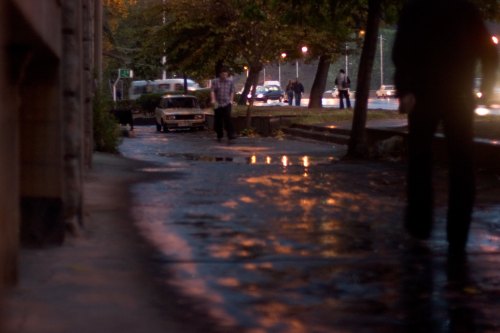 A blurred street scene at dusk, with people walking along a wet sidewalk and cars in the background.