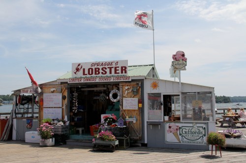 A seafood shack named "Sprague's Lobster" with a flag, flowers, and outdoor seating by the water.