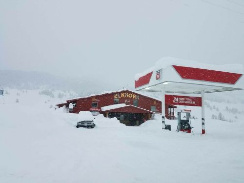A snowy scene featuring a red building and a gas station, both partially buried in deep snow. Visibility is low.