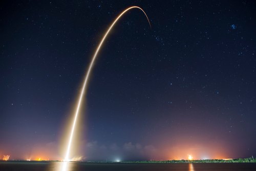 A rocket launches into the night sky, leaving a bright trail against a backdrop of stars and distant lights.