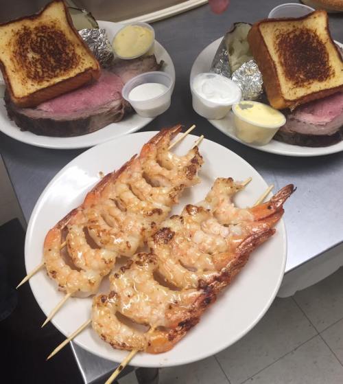 A plate of grilled shrimp skewers with two servings of prime rib and toasted bread in the background.