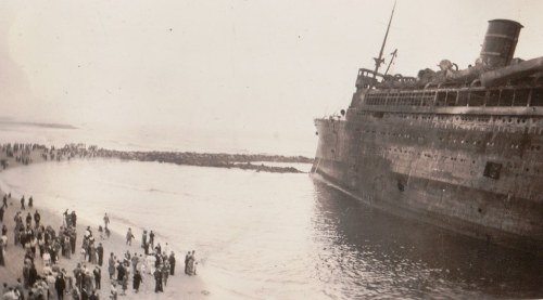 A historic shipwreck on a beach, with a crowd of people gathered on the shore, observing the scene.
