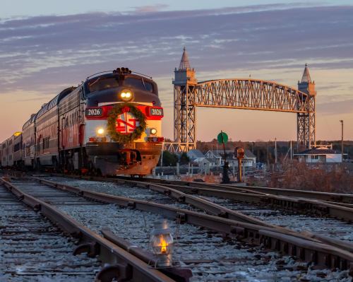 A festive train with a wreath approaches a bridge at sunset, with tracks and a lantern in the foreground.