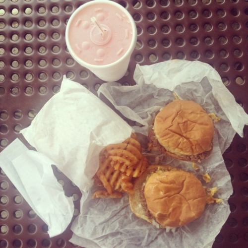 Two burgers wrapped in paper, a side of waffle fries, and a pink drink on a perforated table.
