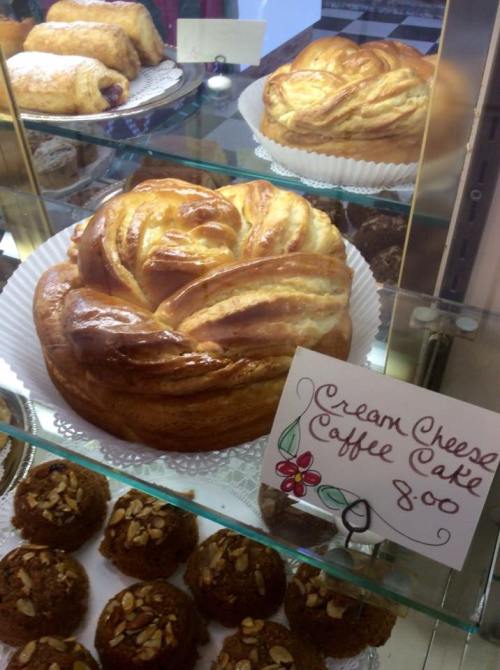 A display case with a cream cheese coffee cake and assorted pastries, including round and nut-topped treats.