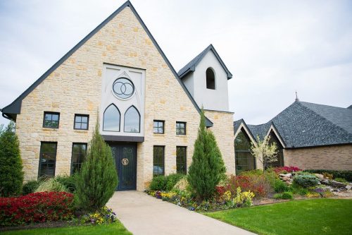 A stone building with large windows, a peaked roof, and landscaped gardens in front.