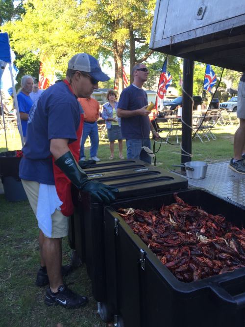 A man in gloves prepares crawfish at an outdoor event, with people and flags in the background.