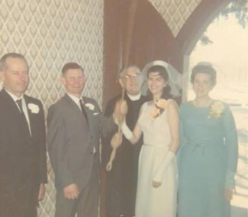 A bride in a white dress holds a bouquet, surrounded by four people, including a priest, in a vintage wedding setting.