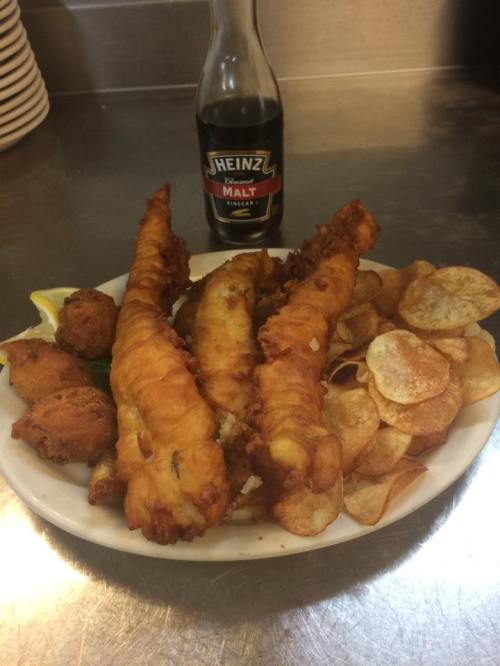A plate of crispy fish fillets, fried potatoes, and a bottle of malt vinegar, garnished with lemon wedges.