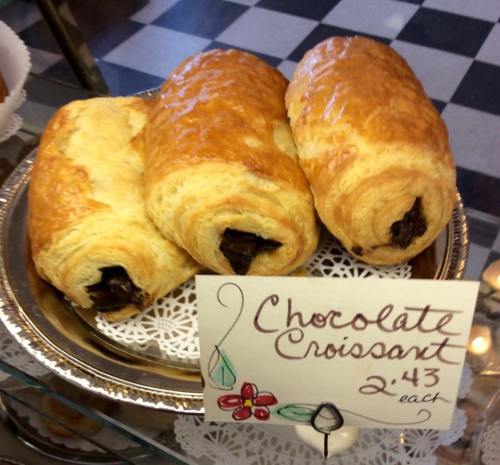Three golden-brown chocolate croissants on a decorative plate, with a sign indicating they cost $2.43 each.