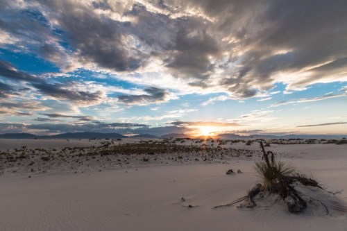 A serene desert landscape at sunset, featuring soft sand, sparse vegetation, and dramatic clouds in the sky.