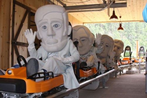 Five large, cartoonish figures with oversized heads wave from roller coaster seats in a wooden building.