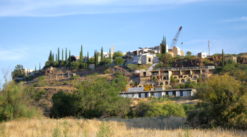A hillside with modern buildings, tall cypress trees, and construction equipment against a clear blue sky.