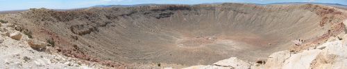 A panoramic view of a large, dry crater with rocky walls and a flat, barren floor under a clear blue sky.