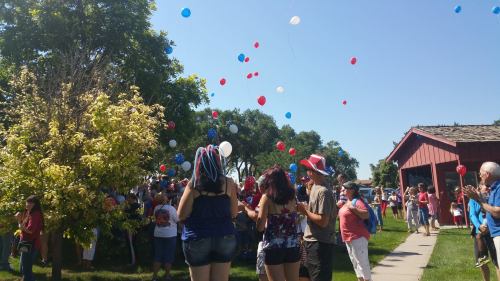 A crowd of people in festive attire releases red, white, and blue balloons into a clear blue sky during a celebration.