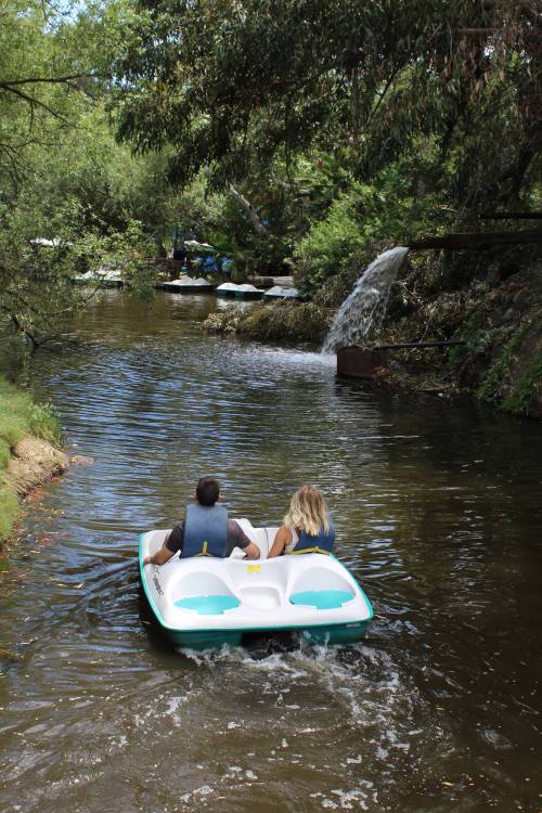 Two people paddle a small boat on a serene, tree-lined waterway with a gentle waterfall in the background.