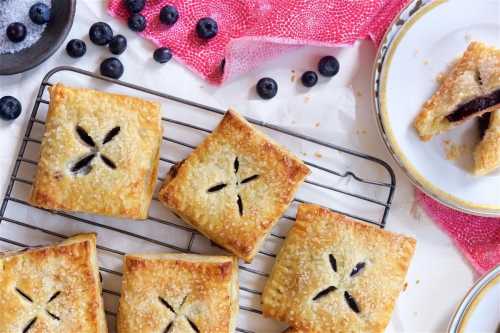 A tray of golden-brown pastries with blueberry filling, sprinkled with sugar, on a cooling rack with fresh blueberries.