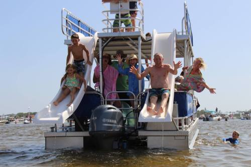 A group of people joyfully sliding down water slides from a boat, with others swimming nearby on a sunny day.
