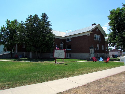 A brick building with a metal roof, surrounded by trees and a lawn, featuring colorful chairs and a sign in front.