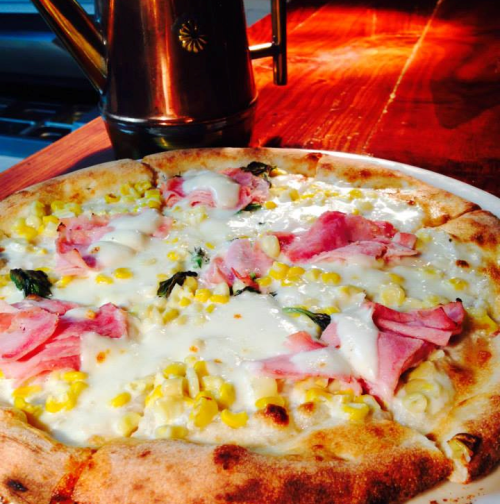 A freshly baked pizza topped with ham, corn, and melted cheese, served on a wooden table with an oil dispenser in the background.